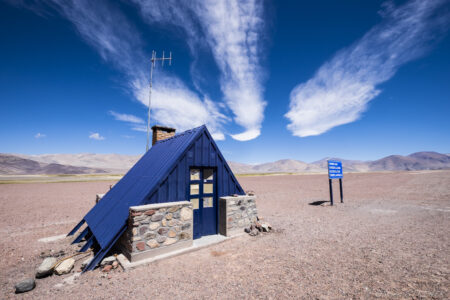 Emergency refuge along the road - Refuge d'urgence le long de la route Desert house in Fiambala, Argentina, with a blue roof and stone foundation, set against a vast, arid.