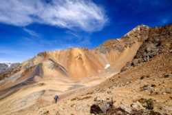 A lone hiker explores a rugged mountain terrain with colorful slopes and a vibrant blue sky, capturing the essence of outdoor adventure photography. Mountain landscape with hiker under blue sky, high-altitude adventure photography.