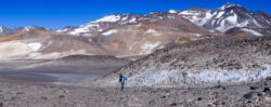 A hiker exploring a volcanic mountain landscape with snow-capped peaks in the background, showcasing rugged terrain and natural beauty. Mountain landscape with hiker in volcanic terrain, high-altitude scenery, and snow-capped peaks.