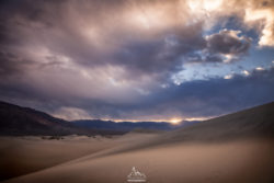 A stunning desert scene featuring rolling sand dunes and a vibrant sunset sky, showcasing natural beauty and atmospheric lighting. Desert landscape with sand dunes under dramatic sky at sunset, captured by Olivier Steiner Photograp.