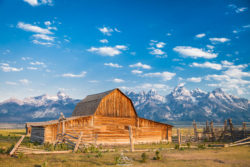 Scenic landscape featuring a rustic wooden barn set against the majestic mountains with a vibrant blue sky overhead. Rustic wooden barn with mountain range in the background, under a bright blue sky.
