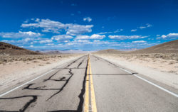 A long, empty highway in a desert setting with cracked pavement and distant mountains, ideal for travel and adventure photography. Open road stretching through a barren desert under a bright blue sky with scattered clouds.