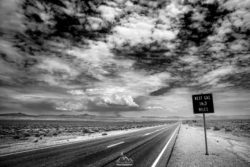 Long, empty desert road with dramatic cloudscape and a sign showing 163 miles to the next gas station. Desert highway with a sign indicating 163 miles to the next gas station.