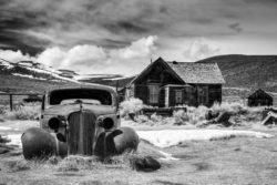 A black and white photo of an abandoned vintage car parked near a rustic wooden house in a rural setting. Old abandoned vintage car in front of a rustic wooden house in a rural landscape.