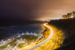 A scenic view of a winding coastal highway illuminated by streetlights, with city buildings in the background and the ocean on the left. Sunset over coastal highway with city lights and ocean view at dusk.