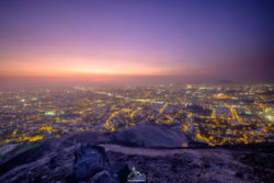 A breathtaking view of a city illuminated at dusk from a mountain summit, highlighting urban lights and natural surroundings. Sunset over a city from a mountain viewpoint, showcasing city lights and scenic landscape.