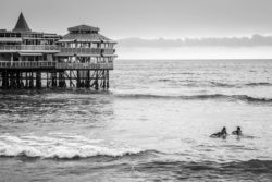 A black and white photo of a pier extending into the ocean with historic buildings, capturing a serene seaside atmosphere. Pier with historic architecture on the beach, black and white ocean scene.