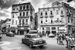 A black and white photo capturing the charm of Havana with classic cars and aged buildings bustling with people. Vintage cars and historic architecture in a lively Havana street scene.