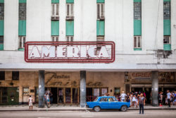 People gather outside the historic America Theater in Havana, Cuba, with a vintage blue car parked in front. Historic America Theater facade with crowd and vintage car in Havana, Cuba.