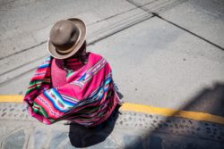 A woman dressed in traditional Peruvian clothing, including a bright, multicolored shawl and hat, sitting on a city sidewalk. Colorful Peruvian woman with traditional attire and hat sitting on the sidewalk.