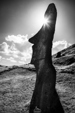 Iconic Moai statue on Easter Island captured in striking black and white, with the sun creating a dramatic sunburst effect. Moai statue on Easter Island with sunburst behind its head, black and white photo.
